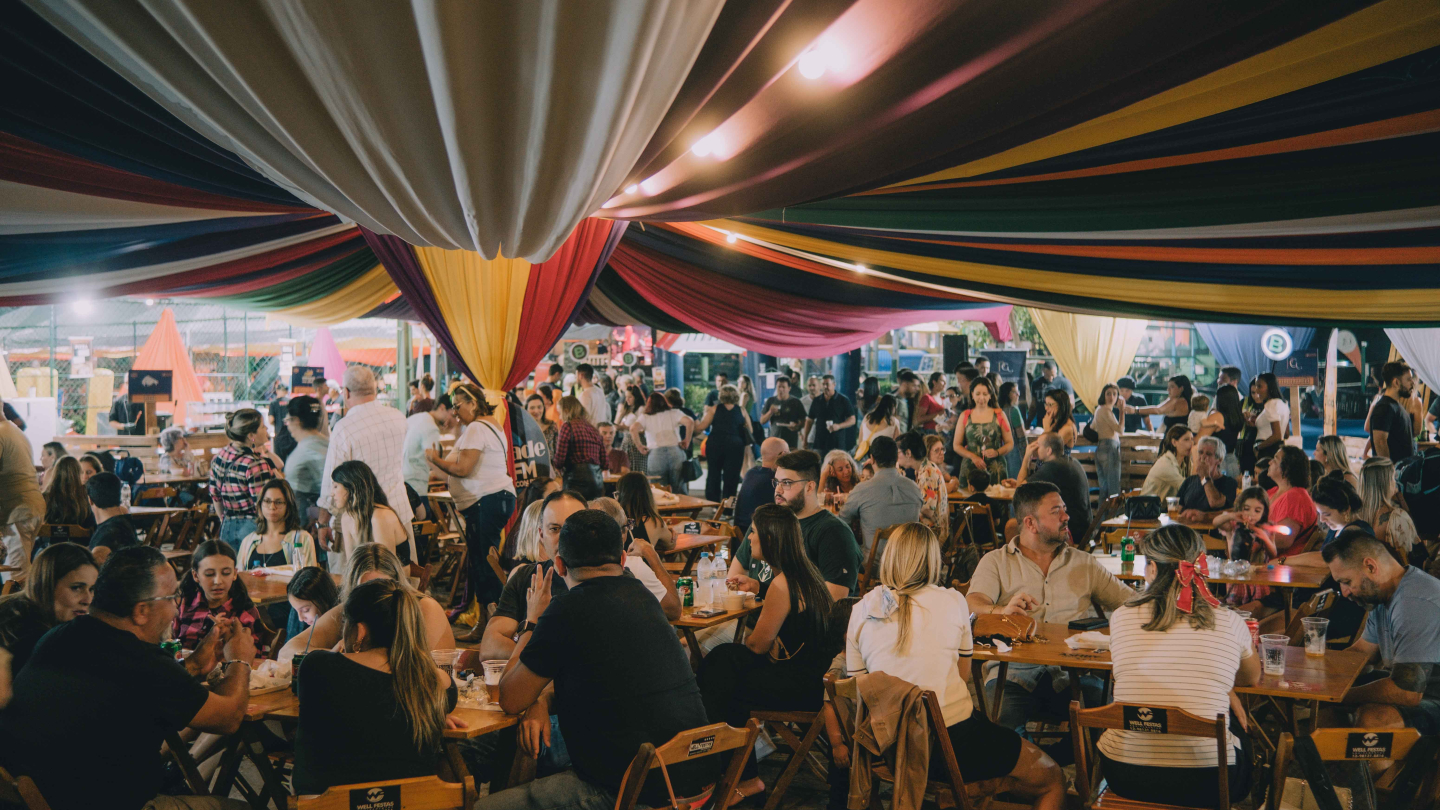 A imagem mostra diversas pessoas reunidas em mesas sob uma grande tenda decorada com tecidos coloridos e luzes suspensas durante um evento festivo