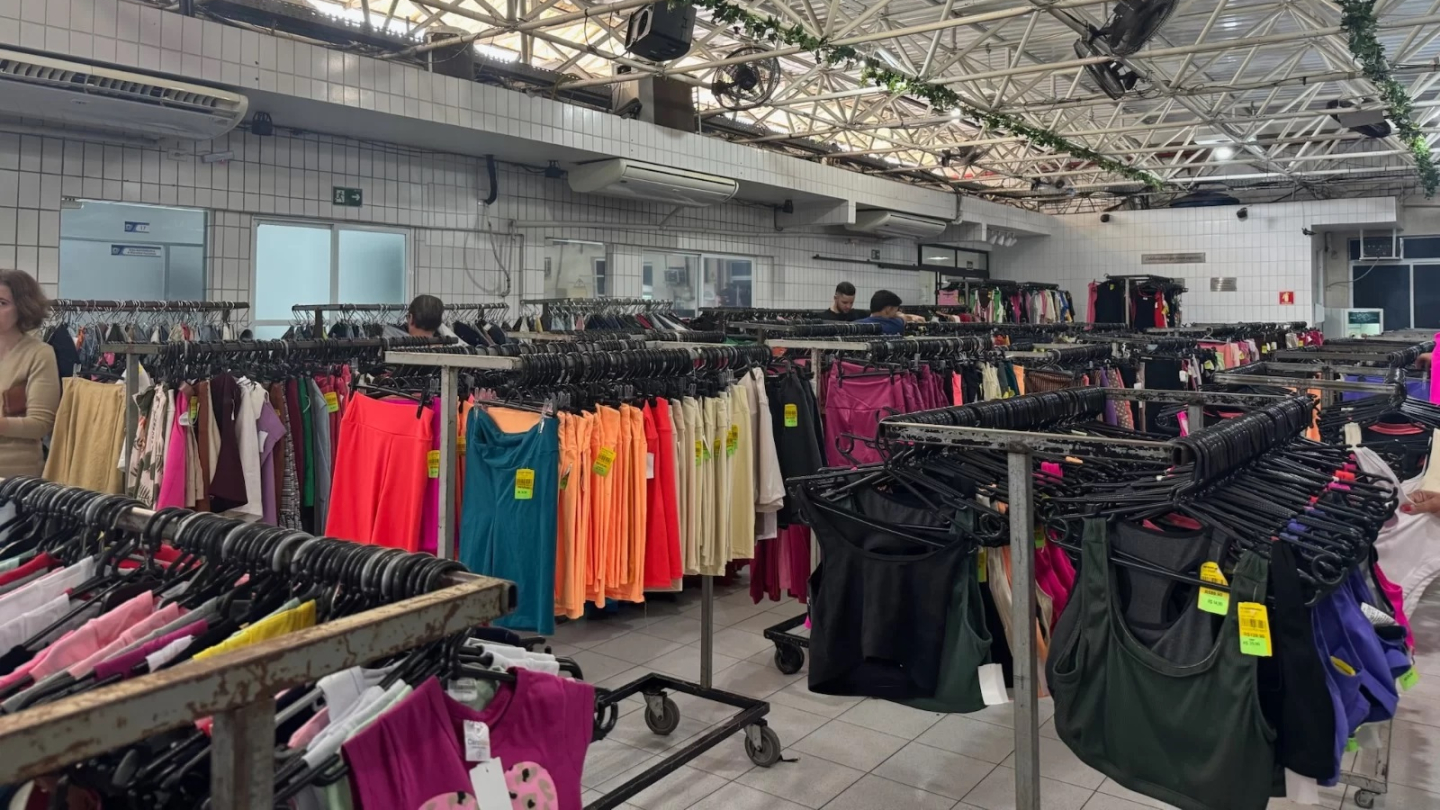 Araras repletas de roupas coloridas e etiquetadas preenchem o amplo salão de um bazar beneficente sob um teto de estrutura metálica
