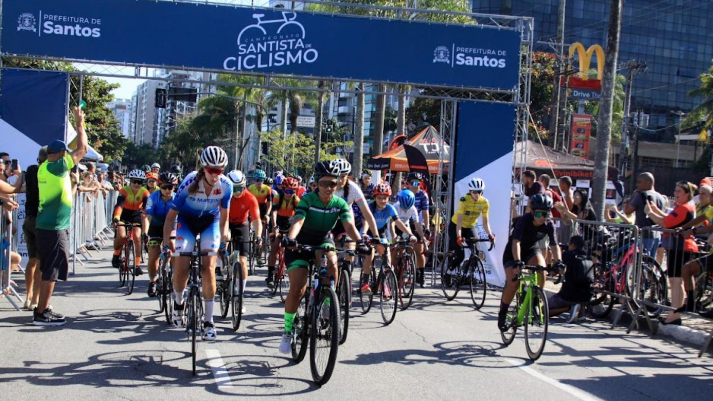 Fotografia de uma competição de ciclismo de rua em um dia ensolarado, mostrando um grupo de ciclistas uniformizados e de capacete largando sob um pórtico azul com a inscrição "Campeonato Santista de Ciclismo"