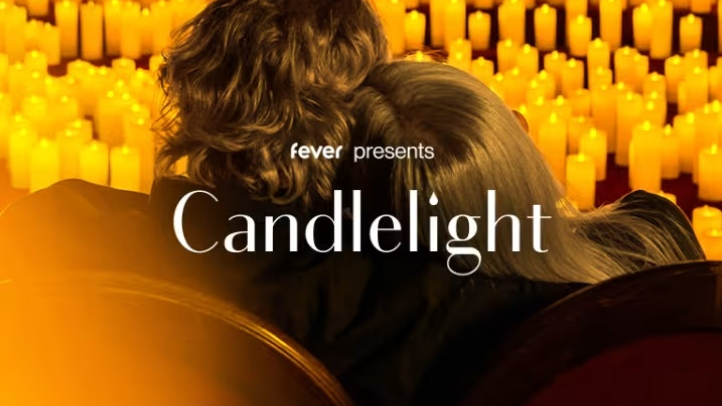 Um casal aparece abraçado de costas, assistindo a um concerto em um ambiente acolhedor iluminado por centenas de velas amarelas