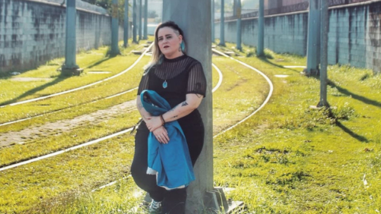 Uma mulher loira com detalhes em azul no cabelo e acessórios posa encostada em um poste metálico entre trilhos de trem sob a luz do sol, segurando um tecido azul