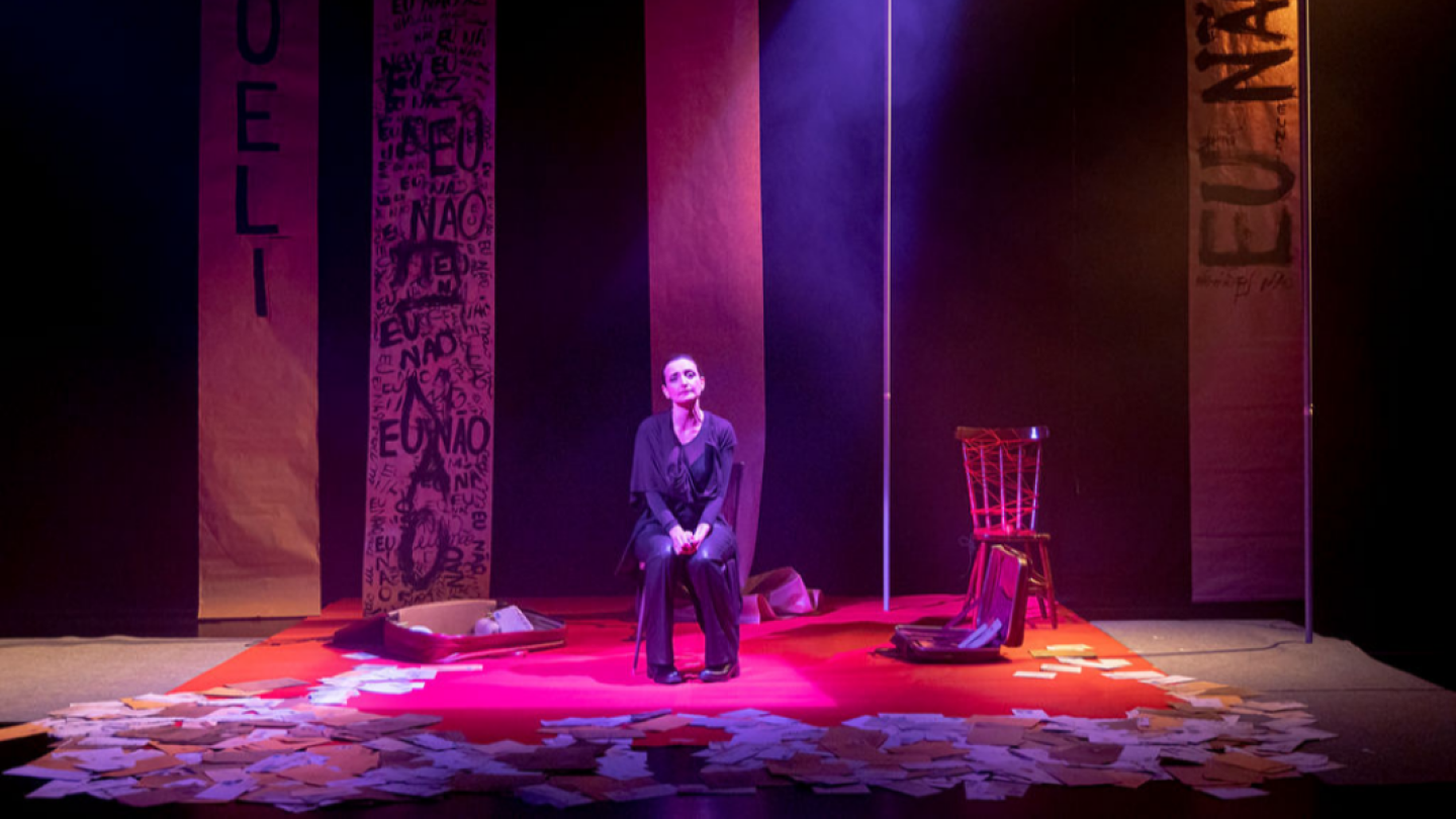 Uma atriz sentada em um palco iluminado por luz roxa é cercada por malas abertas, cartas espalhadas e painéis com inscrições, em uma composição cênica dramática
