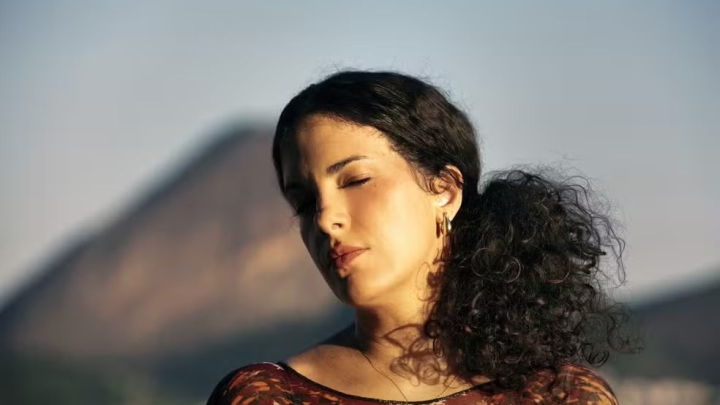 Uma mulher com cabelos cacheados e olhos fechados posa sob a luz do sol, tendo ao fundo uma montanha suavemente desfocada sob um céu claro