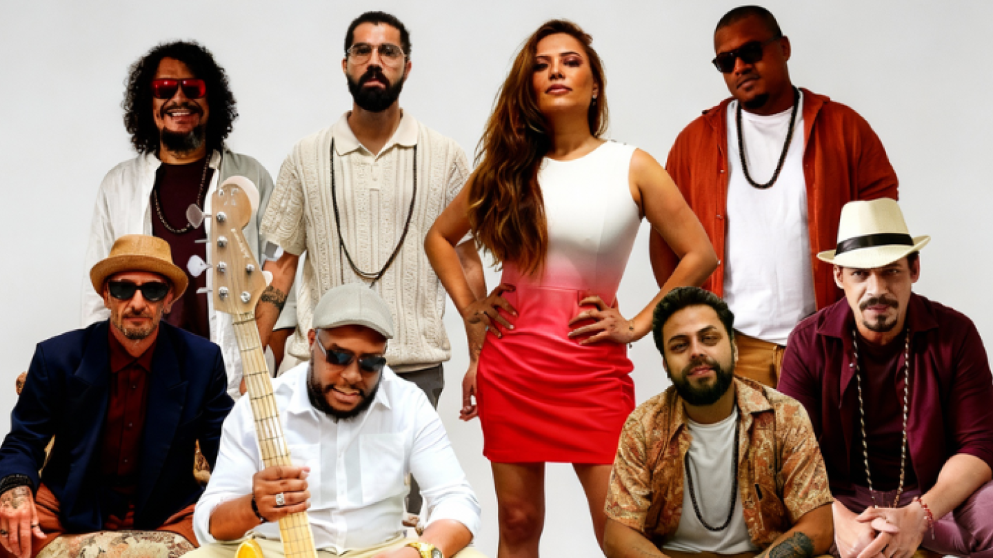 Os integrantes de uma banda musical posam em grupo contra um fundo branco, destacando uma mulher ao centro com um vestido degradê e um baixista em primeiro plano segurando seu instrumento