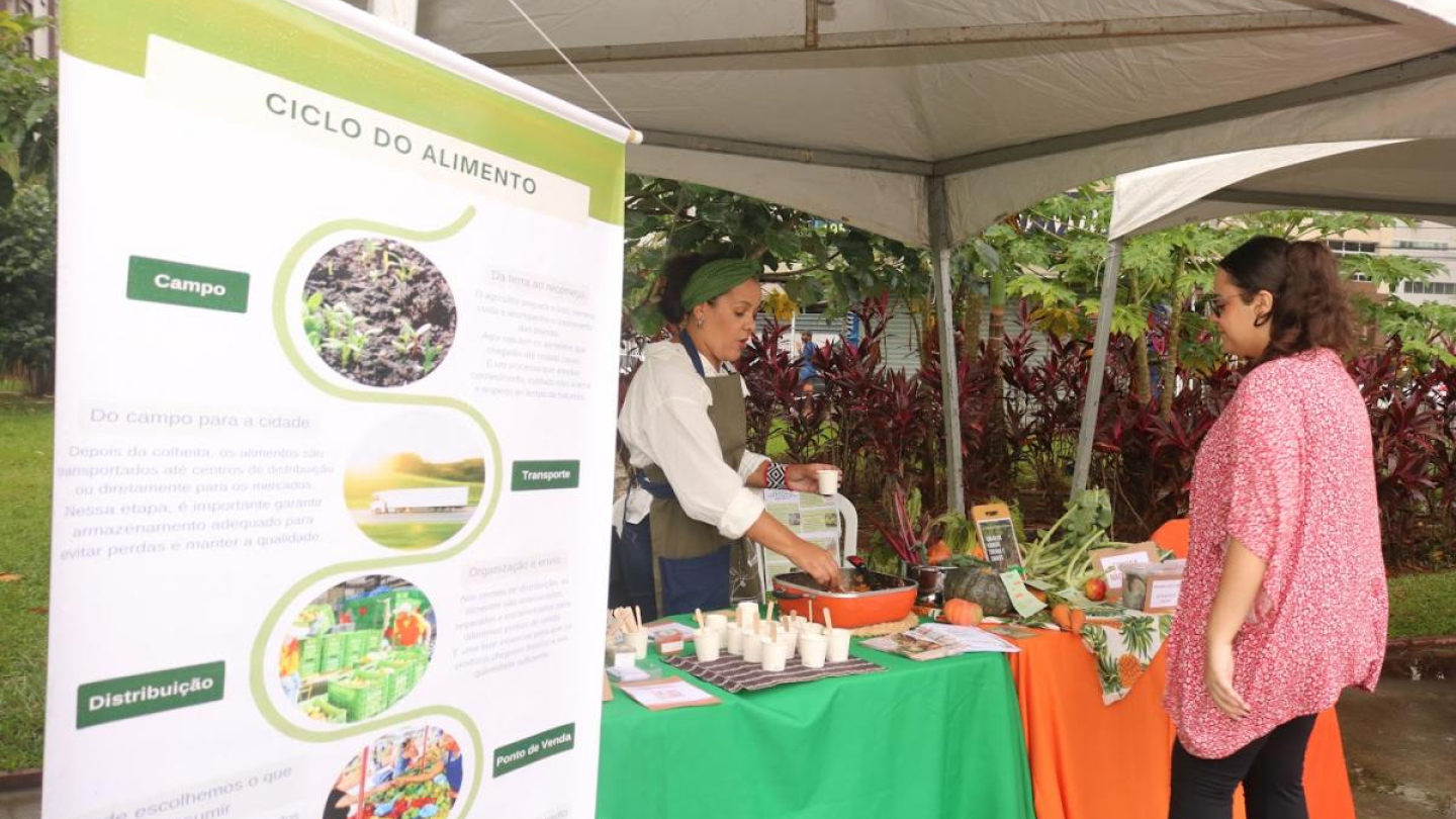 Uma mulher faz demonstração em uma barraca de feira ao ar livre, ao lado de um painel explicativo sobre o "Ciclo do Alimento"