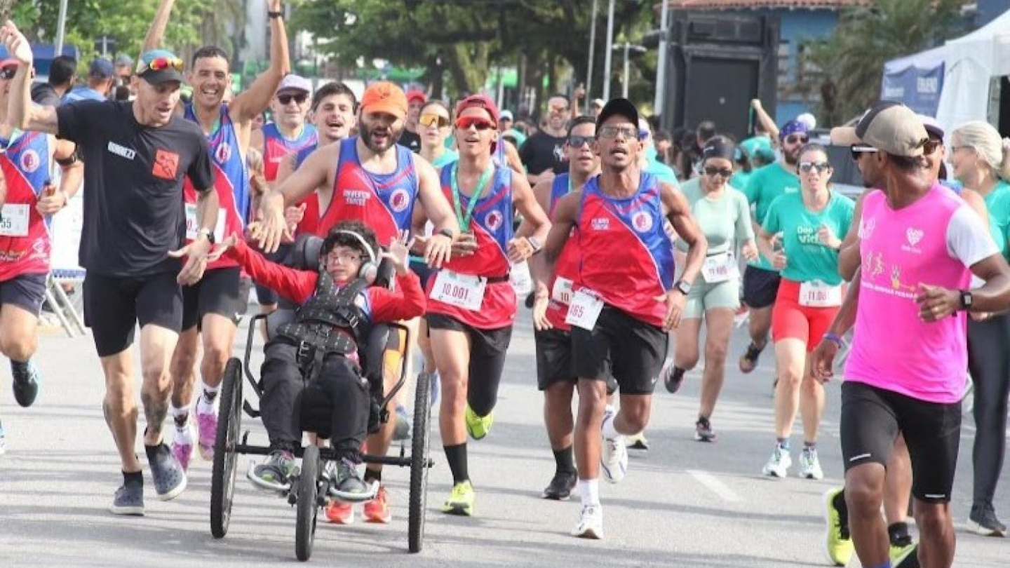 Corredores celebram com entusiasmo durante a prova "Passos para a Inclusão", acompanhando um jovem em uma cadeira de rodas adaptada pelas ruas de Santos
