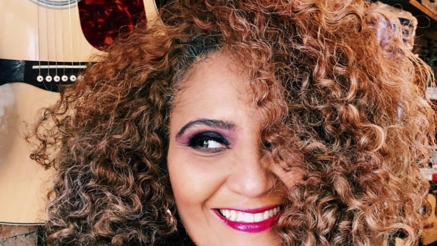 Uma mulher sorridente com cabelos cacheados volumosos e maquiagem marcante olha para o lado, com parte de um violão visível ao fundo