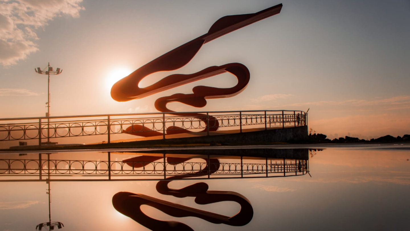 Escultura vermelha de Tomie Ohtake no Emissário Submarino de Santos silhuetada contra o pôr do sol, perfeitamente espelhada em um reflexo na água em primeiro plano