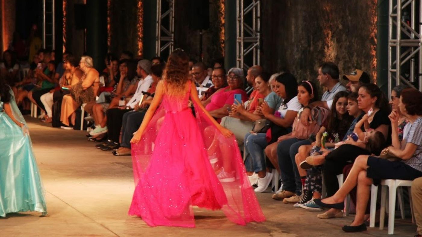 A imagem mostra um desfile de moda em que uma modelo de costas, vestindo um longo vestido rosa vibrante, caminha pela passarela sob os holofotes diante de uma plateia sentada