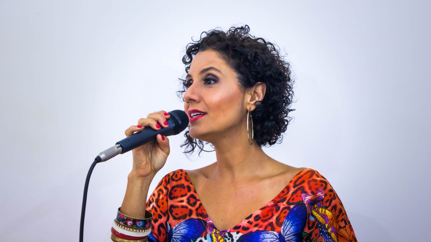 Uma mulher de cabelos curtos e cacheados, usando uma blusa colorida com estampa de borboletas e onça, segura um microfone enquanto fala ou canta contra um fundo branco