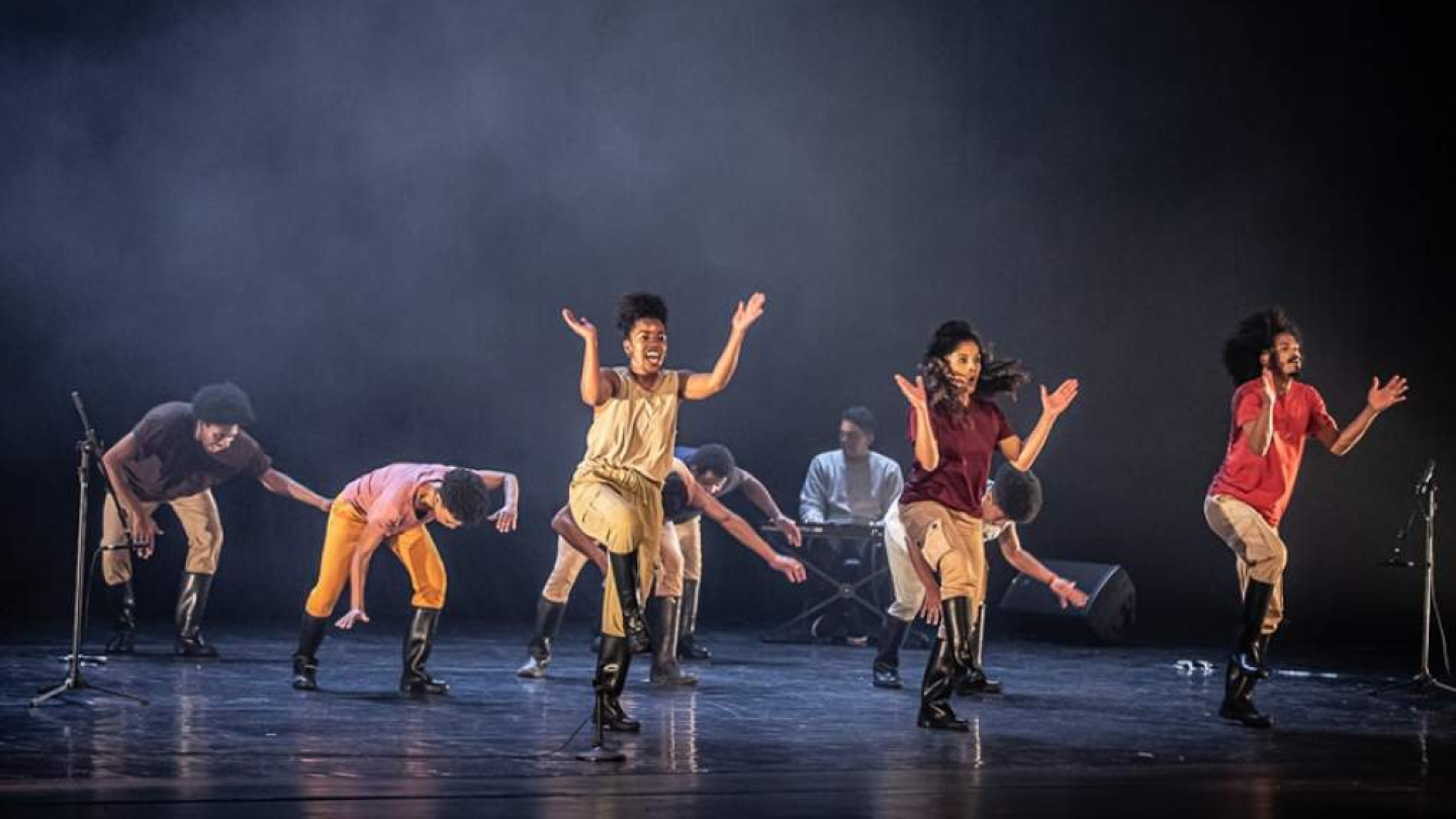 Um grupo de dançarinos negros realiza uma coreografia enérgica de gumboot dance em um palco iluminado, vestindo botas de borracha e roupas coloridas sob uma atmosfera levemente esfumaçada