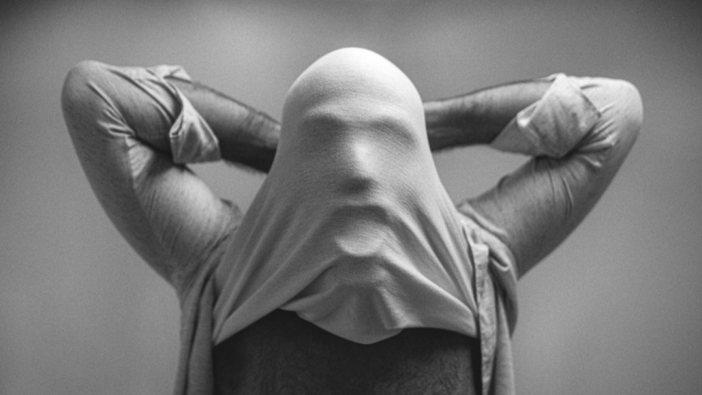 Uma imagem artística em preto e branco mostra uma pessoa com o rosto coberto pelo tecido esticado de sua própria camiseta, criando um contorno facial misterioso e abstrato