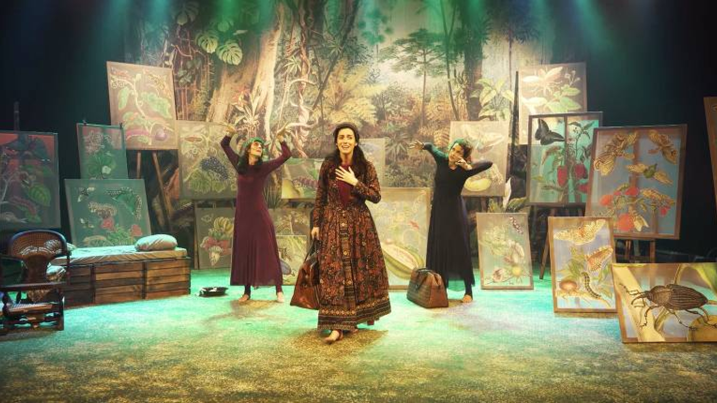 Três atrizes performam em um cenário teatral vibrante que evoca uma floresta, cercadas por diversas telas com ilustrações detalhadas de plantas e insetos.