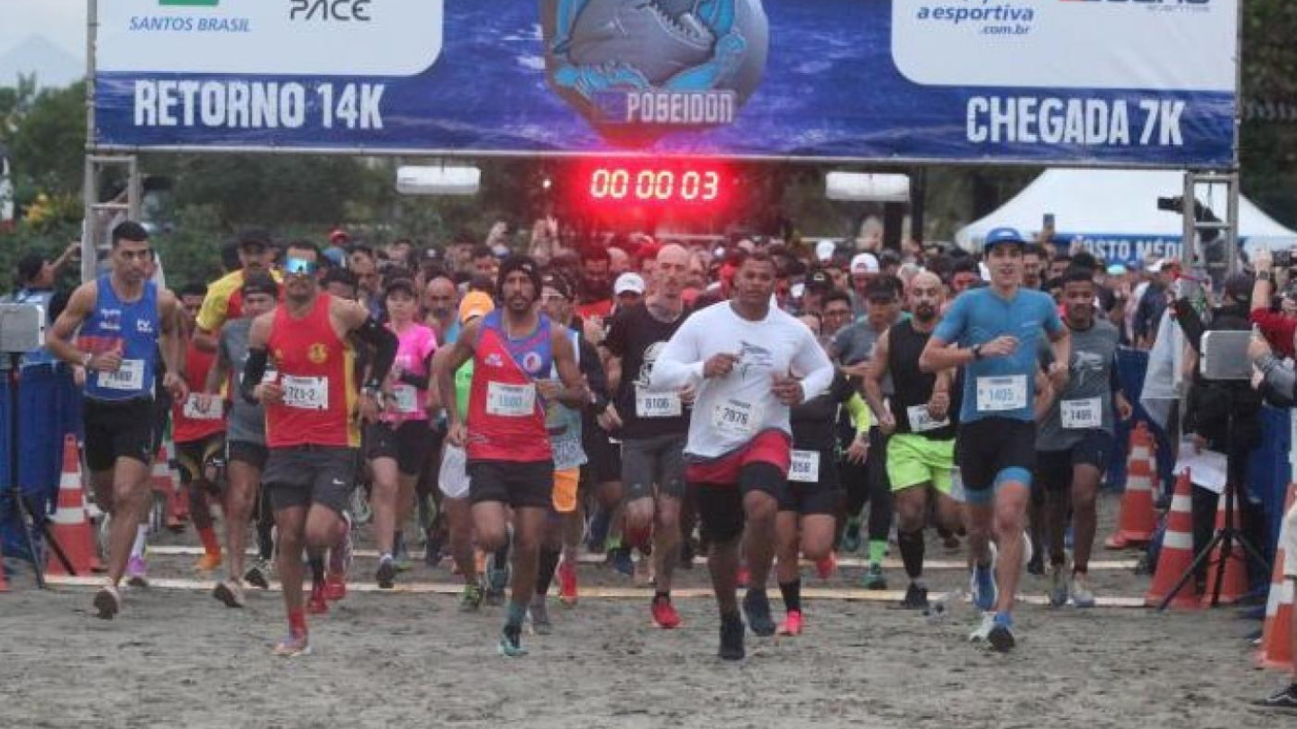 Foto da largada de uma corrida de rua na areia da praia, mostrando diversos atletas passando sob um pórtico de cronometragem do evento "Poseidon"