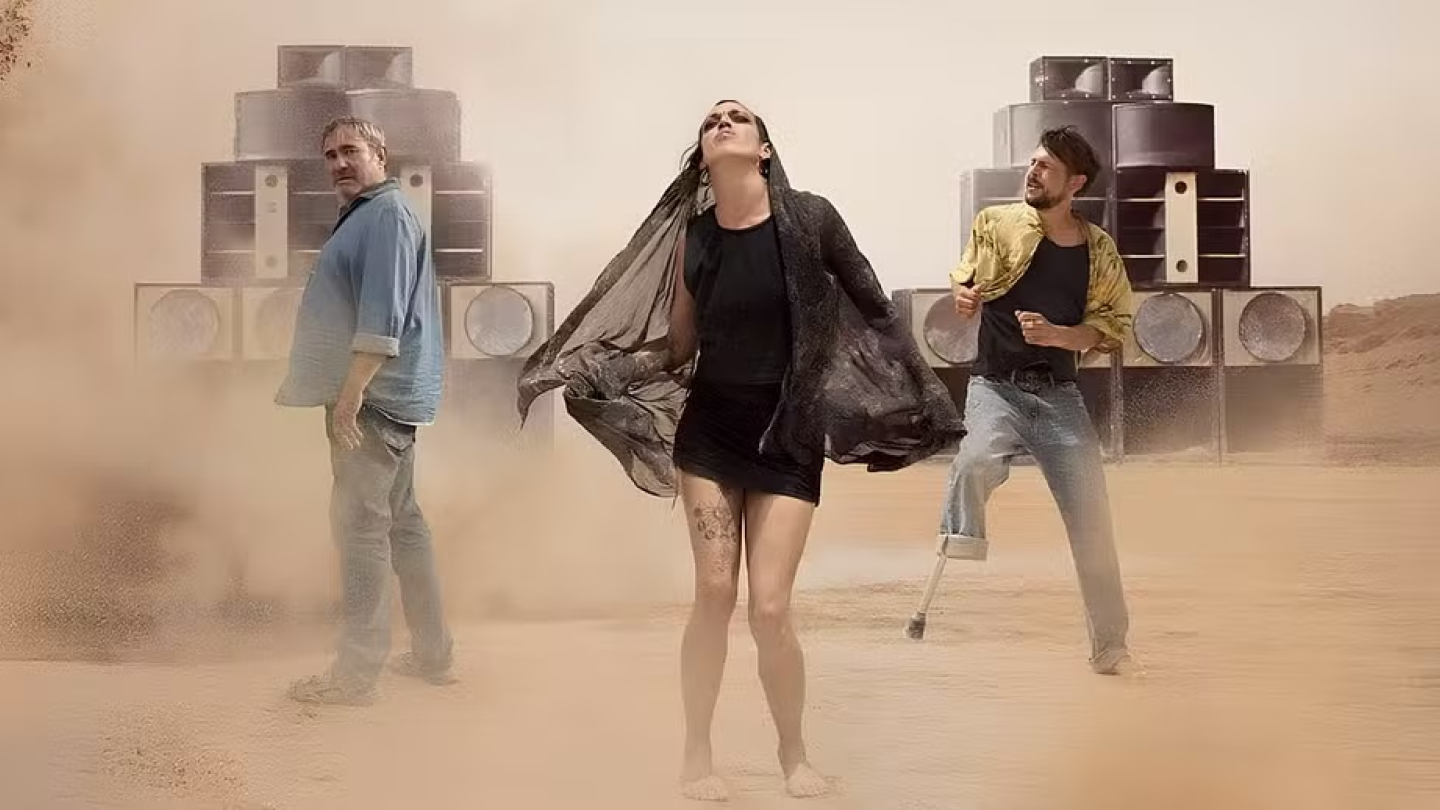 A imagem retrata três pessoas em uma performance expressiva em um ambiente desértico e empoeirado, posicionadas à frente de grandes caixas de som empilhadas