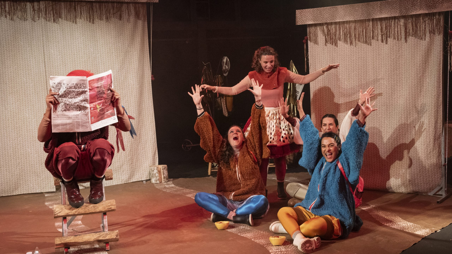 Atores caracterizados no palco de uma peça teatral, onde um deles lê um jornal sentado em um pequeno poleiro de madeira, enquanto os outros expressam entusiasmo e surpresa