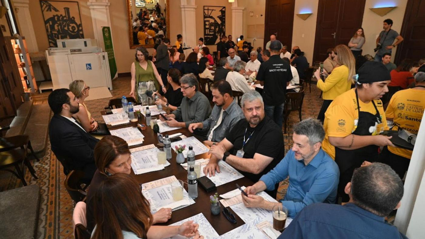 Diversas pessoas estão reunidas e sentadas ao redor de mesas compridas em um amplo restaurante com arquitetura clássica