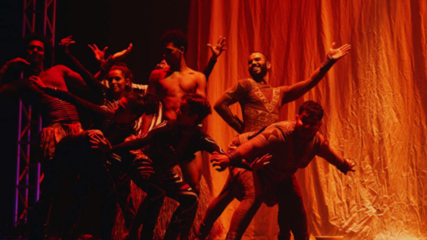 Um grupo de bailarinos realiza uma performance coreografada no palco sob uma iluminação cênica avermelhada e em frente a uma grande cortina de tecido