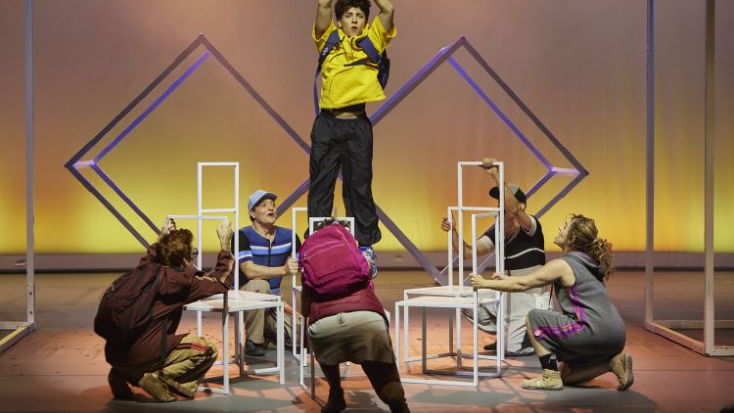 Atores no palco de uma peça teatral, onde um jovem de camisa amarela e mochila equilibra-se em pé sobre estruturas cúbicas brancas, amparado por outros integrantes do elenco ao seu redor
