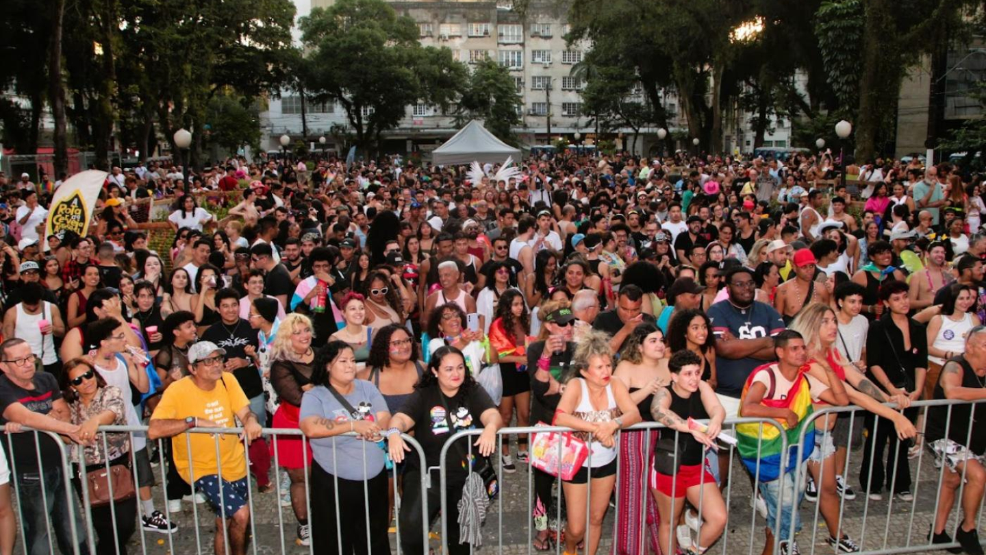 Uma grande multidão se reúne em uma praça arborizada durante um evento da Parada LGBT+, com pessoas concentradas atrás de grades de proteção metálicas