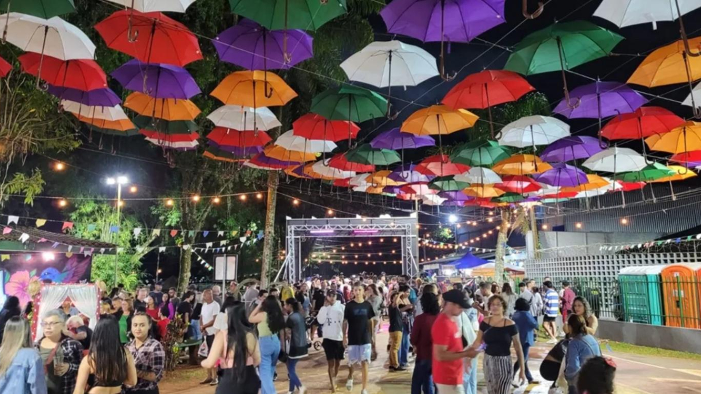Uma grande multidão passeia por uma festa ao ar livre decorada com bandeirinhas, luzes suspensas e muitos guarda-chuvas coloridos no teto