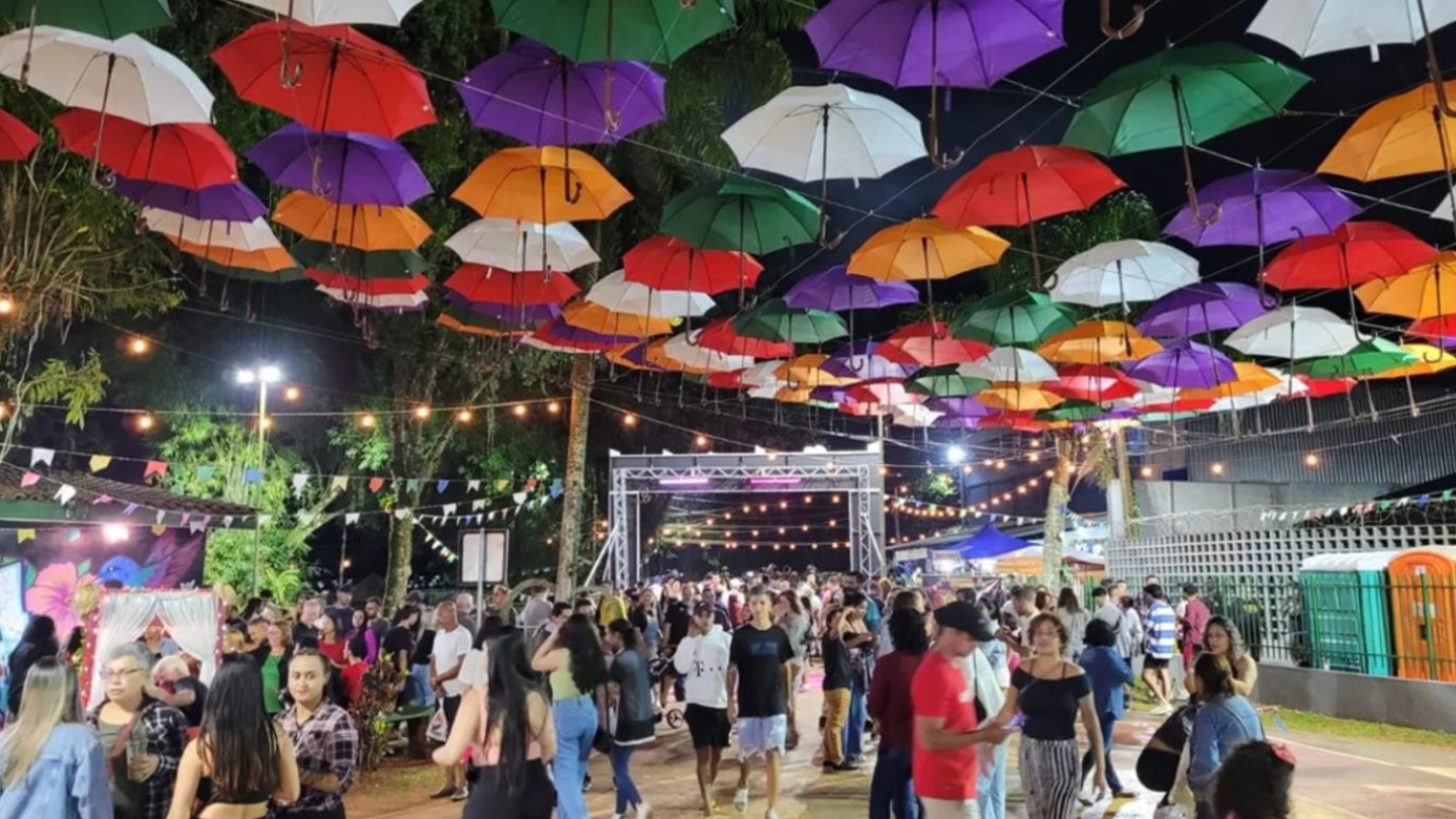 Uma grande multidão passeia por uma festa ao ar livre decorada com bandeirinhas, luzes suspensas e muitos guarda-chuvas coloridos no teto