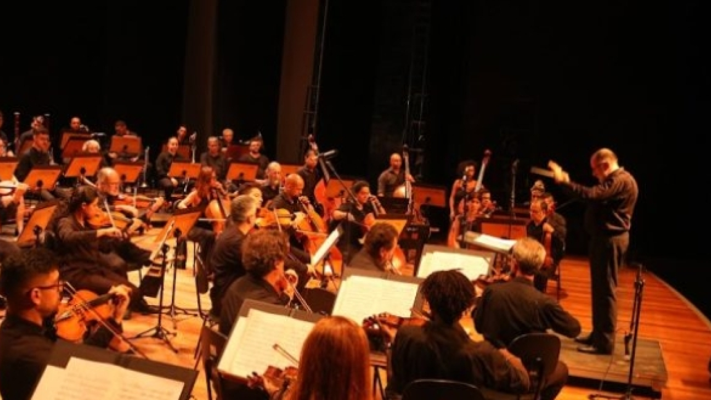 Fotografia de uma orquestra sinfônica durante uma apresentação em teatro. Dezenas de músicos ocupam o palco tocando instrumentos de cordas, sopros e percussão, enquanto um maestro rege o conjunto à frente. A iluminação quente em tons alaranjados destaca os músicos, as estantes de partituras e a atmosfera solene do espetáculo.