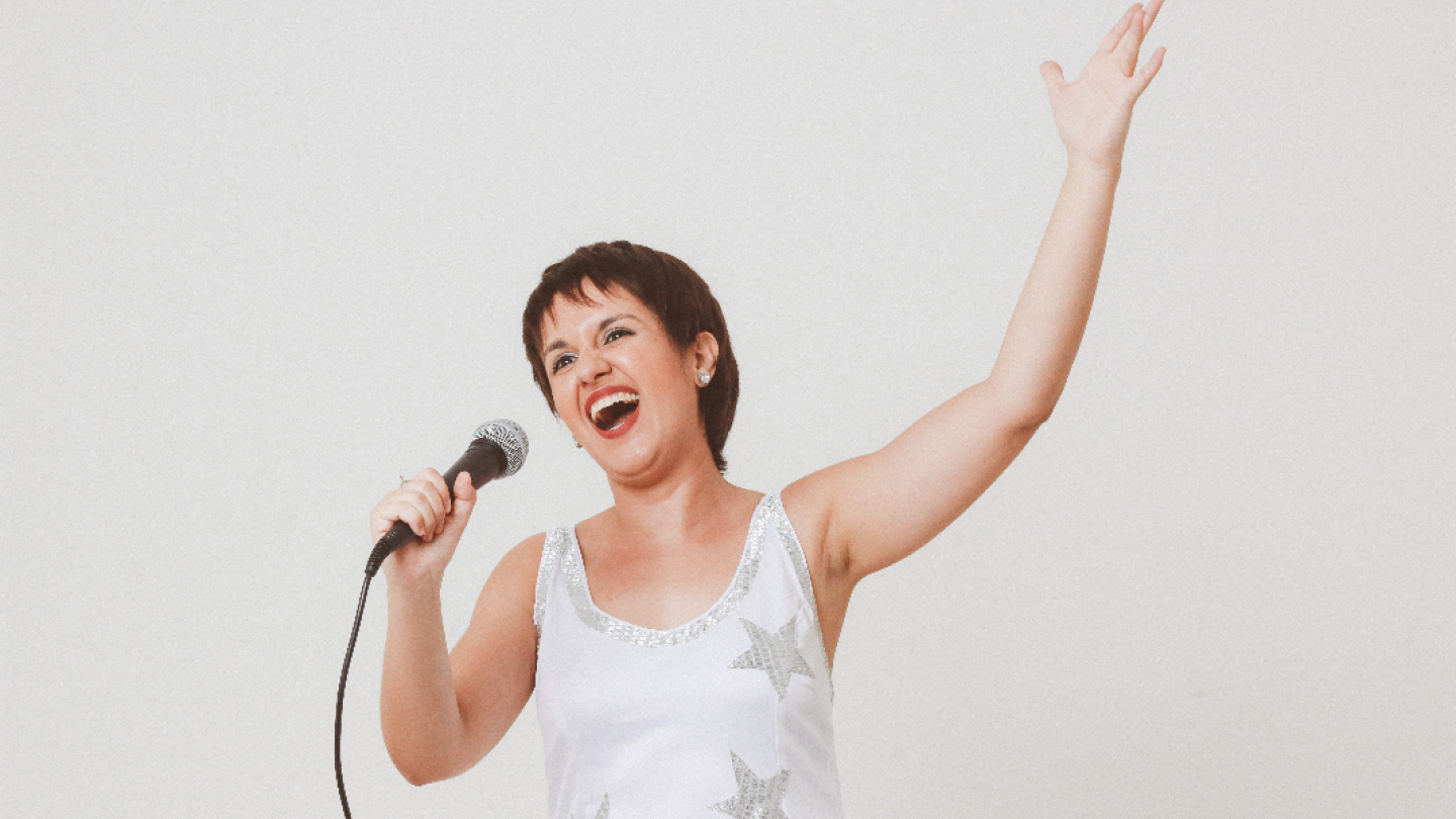 Fotografia de uma cantora durante uma apresentação, segurando um microfone enquanto canta com um sorriso aberto e expressão de entusiasmo. Com o braço direito erguido, ela transmite energia e emoção, como se estivesse interagindo com o público. Veste uma blusa branca com detalhes em brilho no decote e aplicações de estrelas prateadas. O fundo claro e sem elementos de distração destaca a artista e reforça o foco em sua performance vibrante.