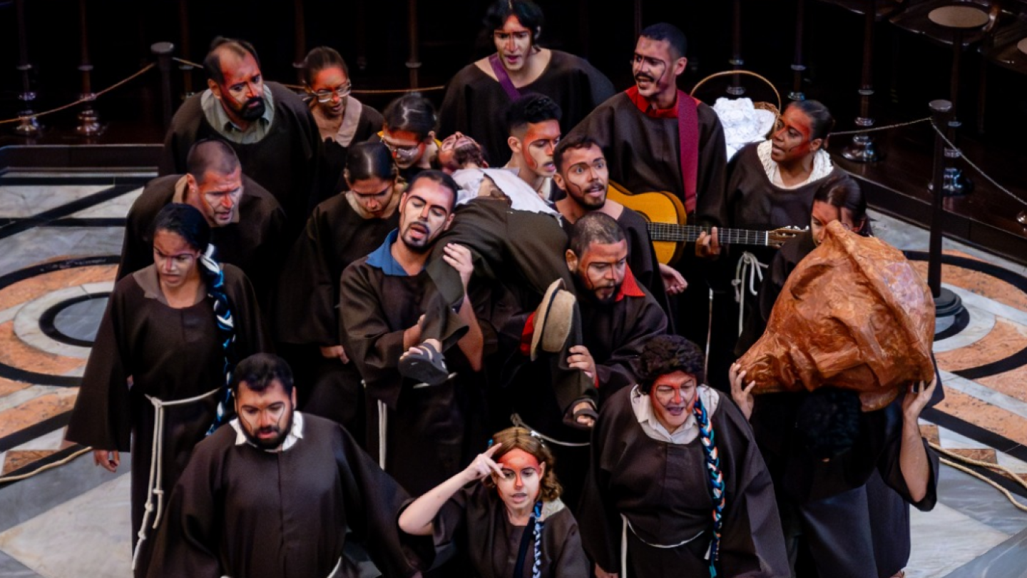 Elenco numeroso ocupa o palco em uma cena coletiva, com atores usando figurinos em tons escuros e maquiagem cênica. Alguns carregam um personagem e objetos de cena, enquanto um músico toca violão ao fundo, criando uma composição intensa e dinâmica que remete a uma procissão ou cortejo teatral.