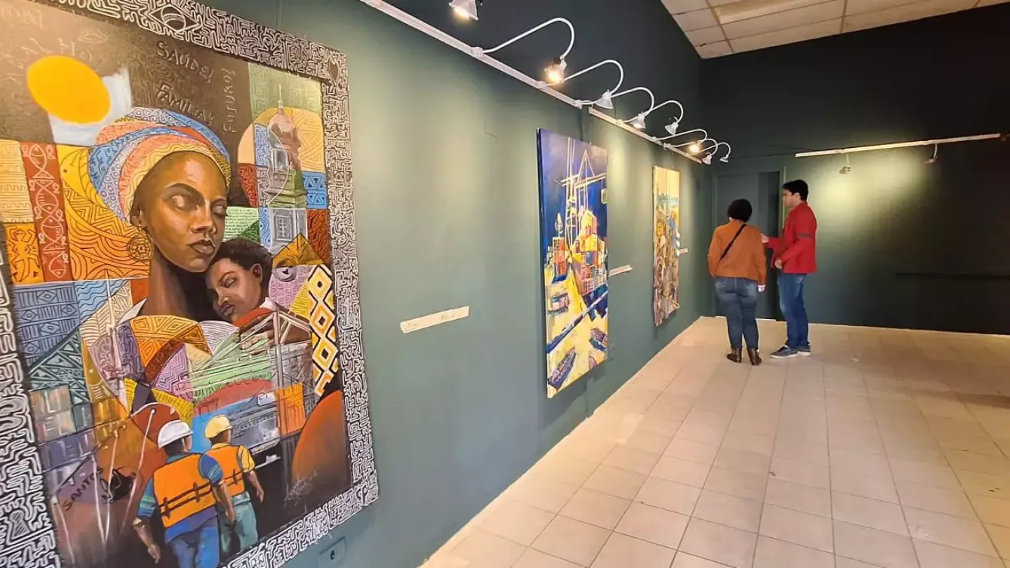 Fotografia do interior de uma galeria de arte com paredes escuras e iluminação direcionada para as obras. Em primeiro plano, destaca-se uma grande pintura colorida retratando uma mulher com uma criança no colo, cercadas por elementos que remetem ao Porto de Santos e à cultura local. Ao longo da parede, outras telas estão expostas, enquanto dois visitantes observam as obras ao fundo do espaço.