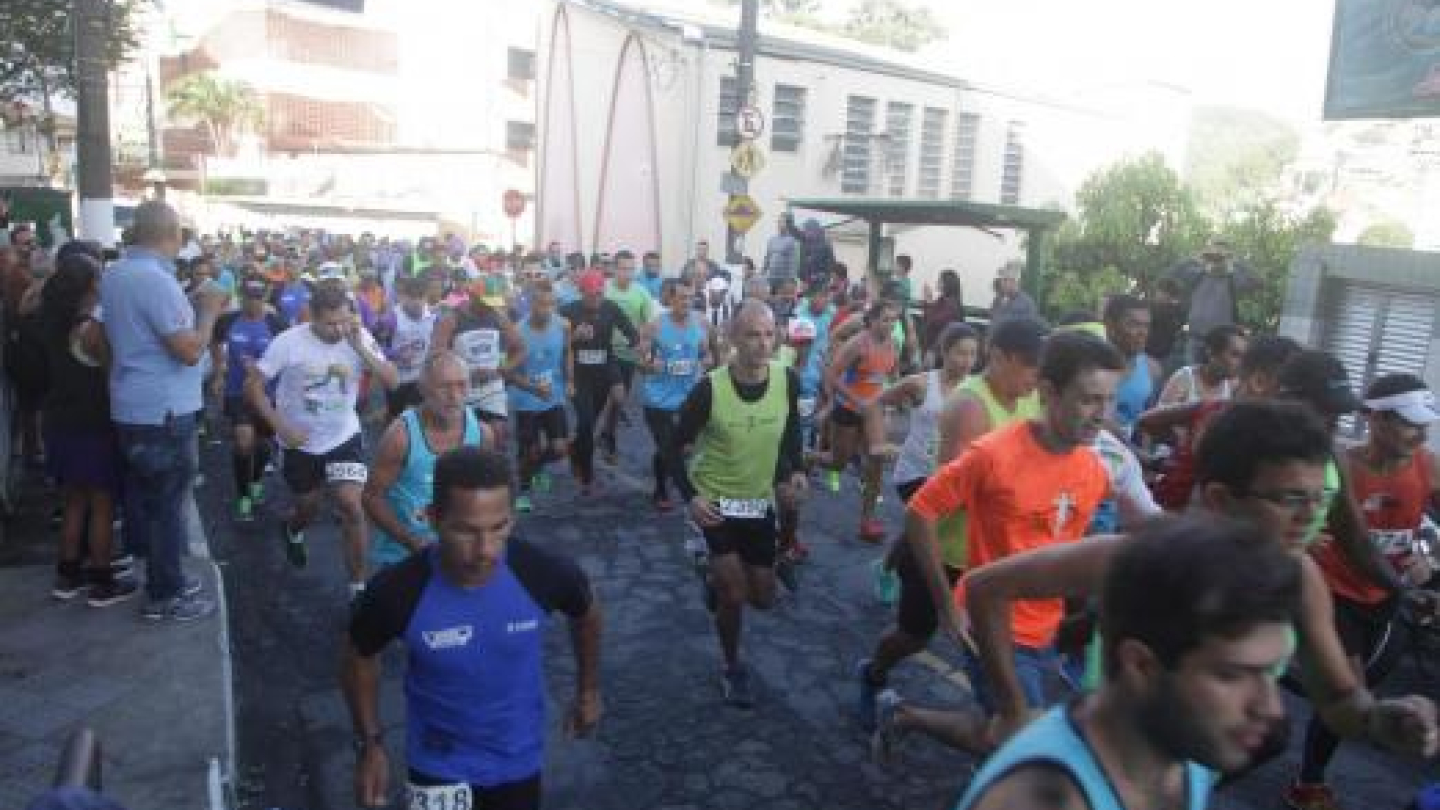 A imagem mostra a largada de uma corrida de rua realizada em uma via dos morros de Santos. Dezenas de corredores, usando camisetas esportivas coloridas e números de peito, seguem pelo percurso enquanto espectadores acompanham a prova nas calçadas. Ao fundo, aparecem imóveis da região e a paisagem urbana característica dos morros, evidenciando o clima de competição e participação comunitária.