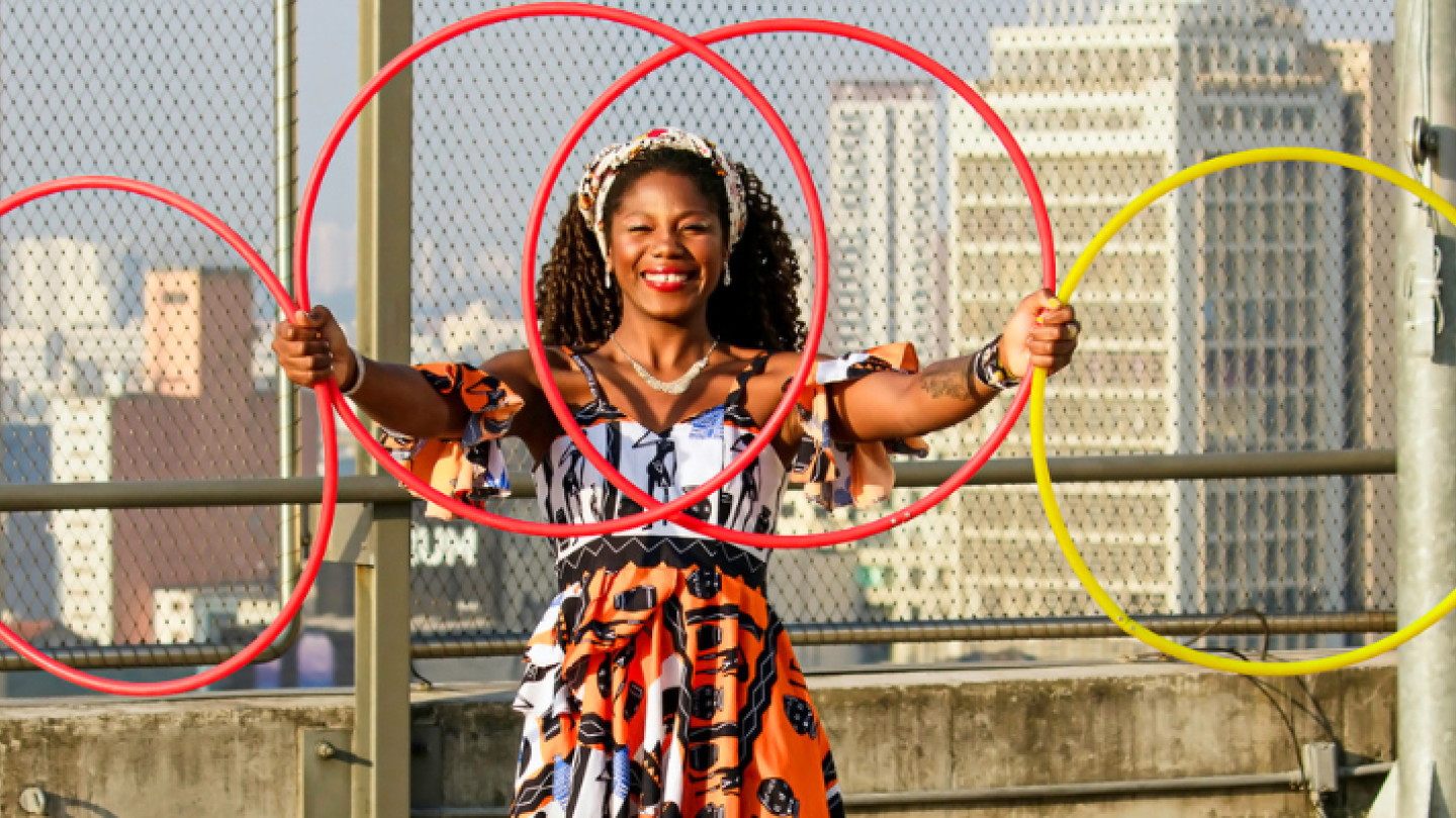 Fotografia de uma artista sorridente segurando quatro bambolês coloridos — vermelhos e amarelo — formando um conjunto de círculos à sua frente. Ela veste um vestido estampado em tons de laranja, preto e branco, além de acessórios no cabelo e um colar. Ao fundo, é possível ver uma tela de proteção e a paisagem urbana de Santos, com prédios em destaque. A composição transmite leveza, movimento e alegria, remetendo a atividades circenses, artísticas e de expressão corporal.